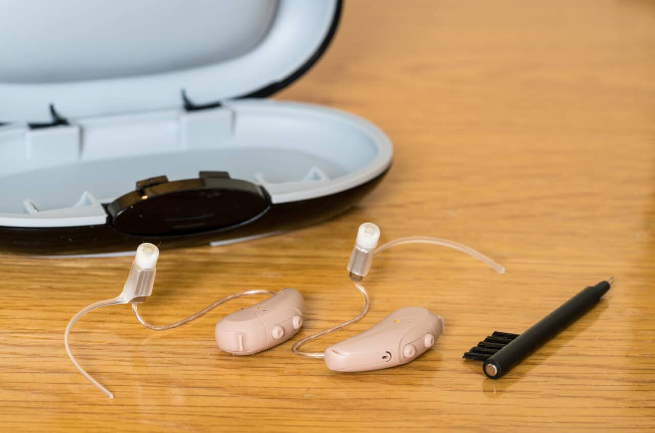Small hearing aids next to a case and brush on wooden bedside dressing table.