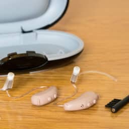 Small hearing aids next to a case and brush on wooden bedside dressing table.