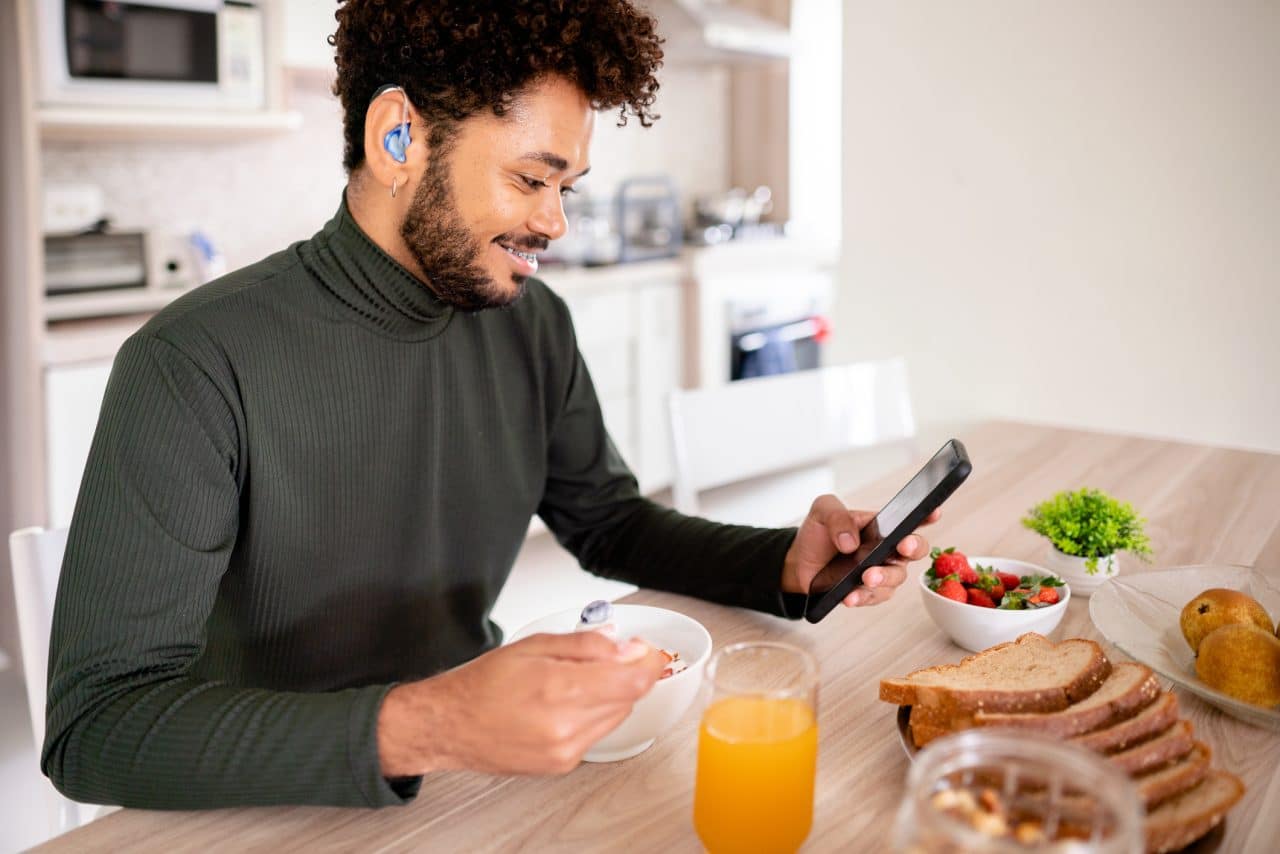Young man with a hearing aid checking his phone over breakfast.